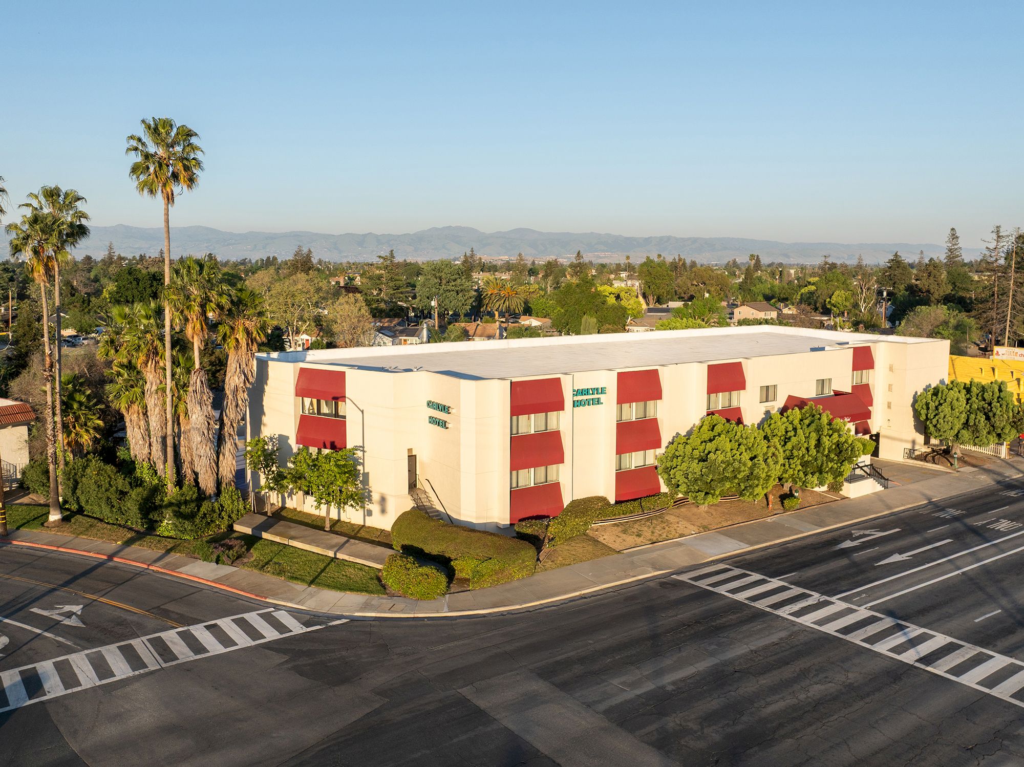 An airy, two-story white building with red accents sits at a street intersection, surrounded by palm trees and a clear sky, in a suburban setting.