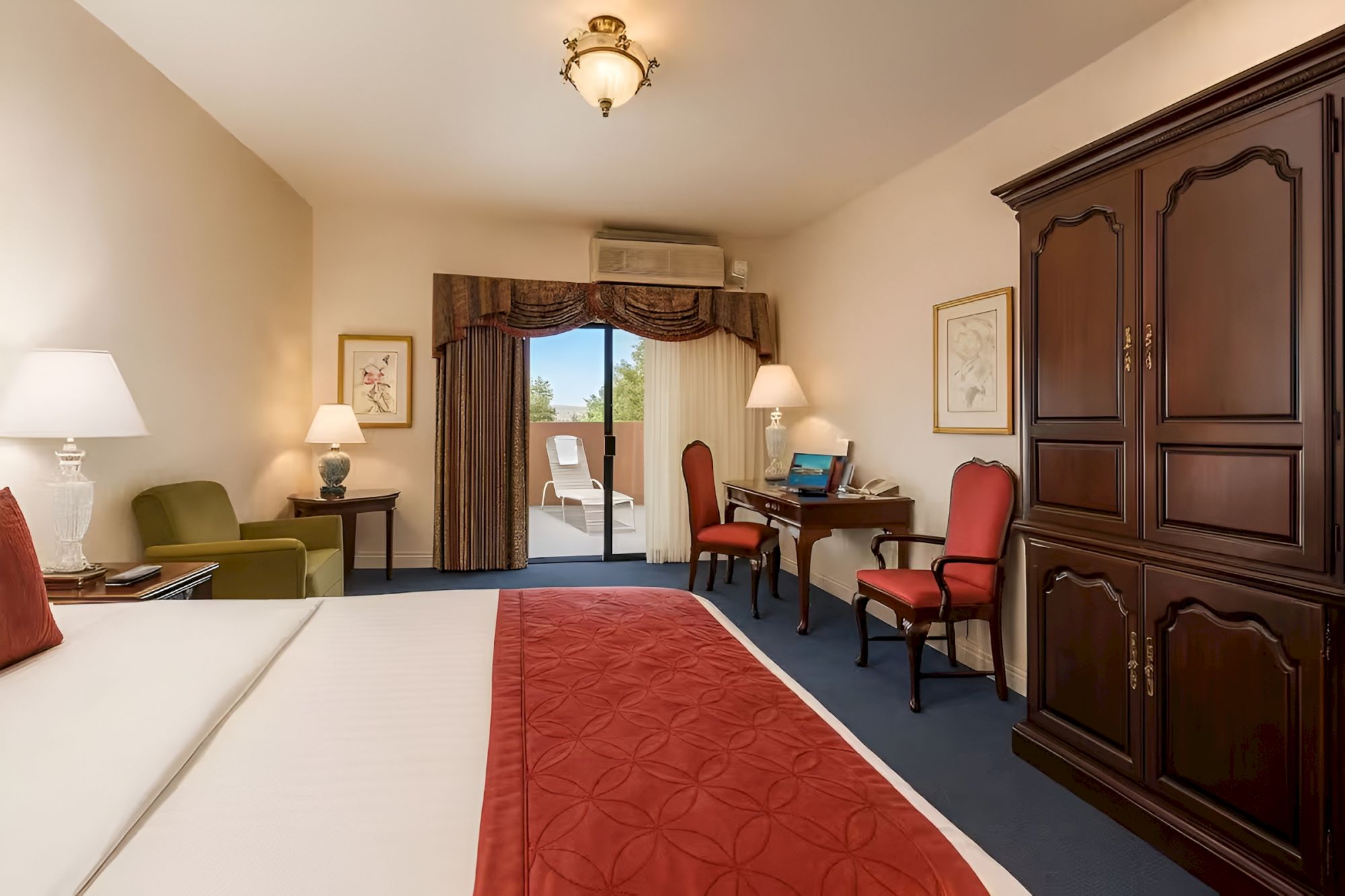 A hotel room with a large bed, red accents, seating area, desk, wardrobe, and a balcony door leading outside. It looks cozy and well-lit.