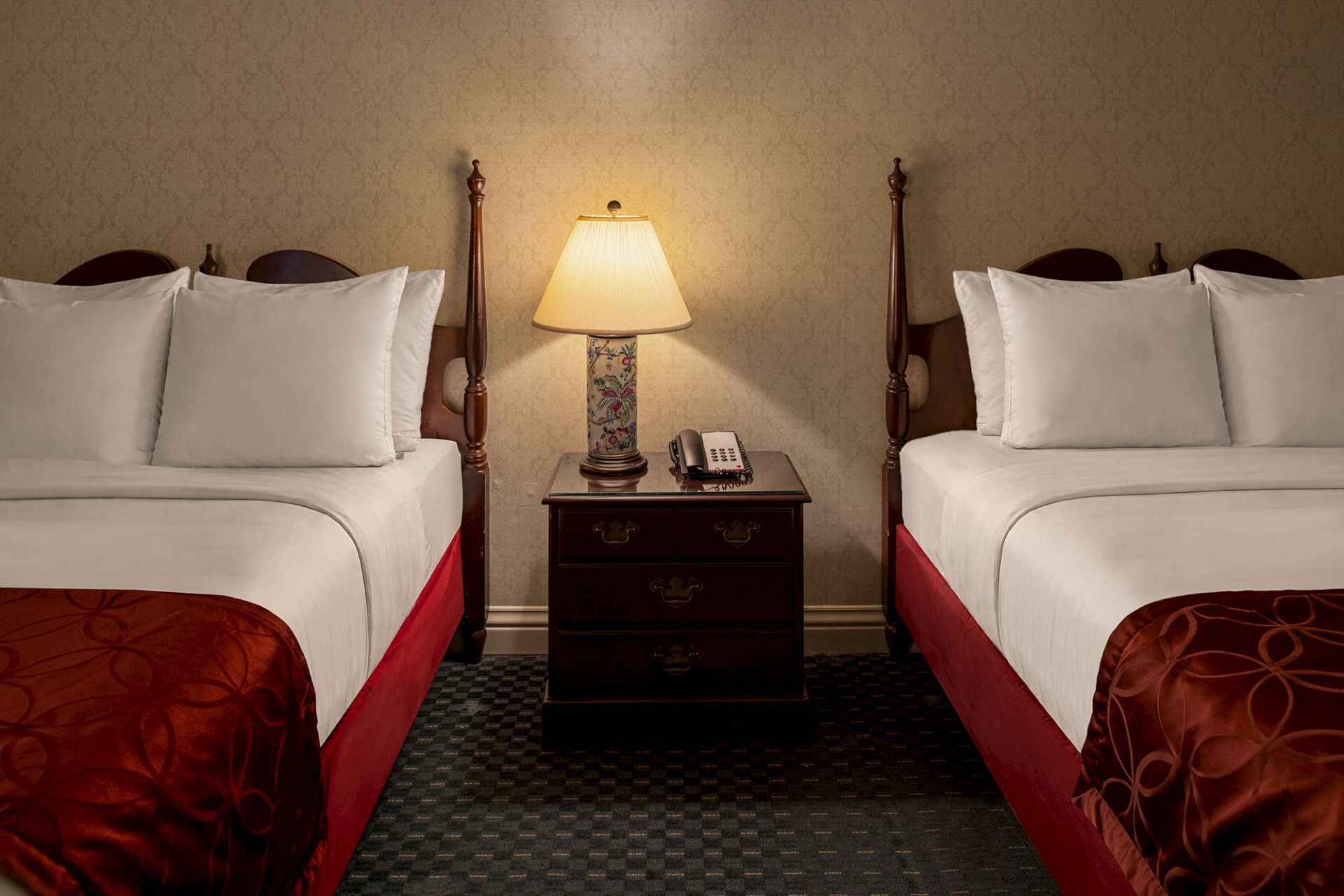 Two hotel beds with white pillows, a central wooden nightstand, and a lit table lamp on a patterned carpet.