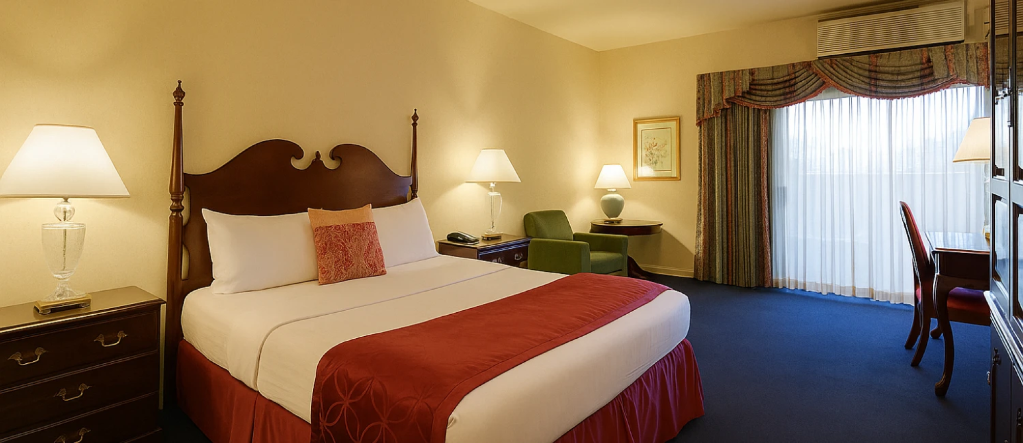 A hotel room with a large four-poster bed, red accents, two bedside lamps, and a sitting area by a window.