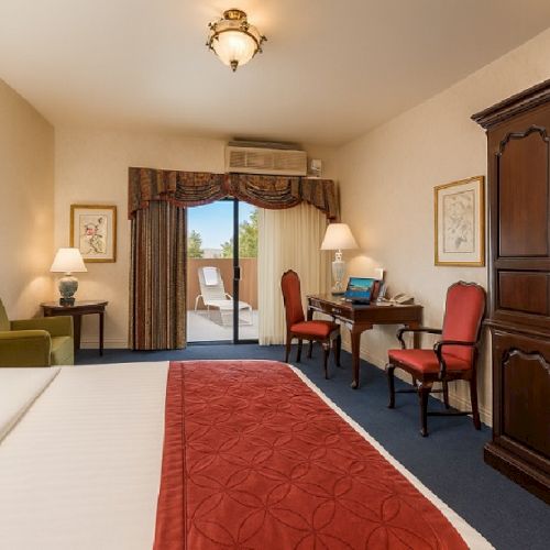 A tidy hotel room with a large bed, red accents, desk and chairs, a wooden armoire, lamps, and a sliding door to a balcony.