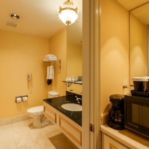 A clean hotel bathroom with beige walls, a double sink, a toilet, a woven basket, towels, and a small coffee station near a decorative window.