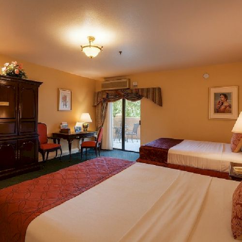 A cozy hotel room with two double beds, warm beige walls, a desk with chair, a wardrobe, a sofa, and a balcony door, lit by ceiling and table lamps.