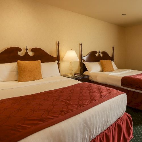 Two double beds in a hotel room with wooden headboards, white linens, red runners, and a nightstand with a lamp in between.