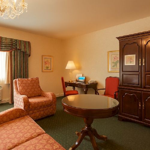 A cozy hotel living area with a floral sofa, matching chairs, a round coffee table, a wooden cabinet, desk with chair, lamp, and window with draped curtains, warm lighting.