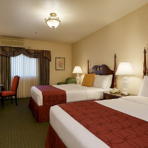 A hotel room with two double beds, warm beige walls, red bed runners, bedside lamps, a dark wood desk, and a window with draped curtains.