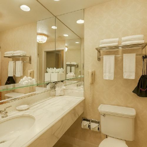 Small hotel bathroom with double sink, large mirror, beige walls, orange shower curtain, towels on rack, trash bag, and toilet, clean and bright.