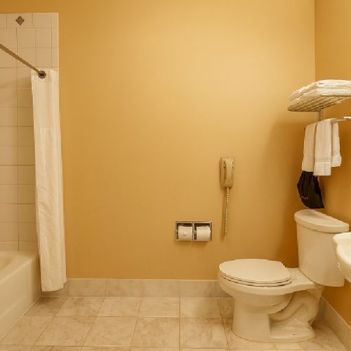 A beige-tiled bathroom with a bathtub and shower on the left, a toilet, a sink, and shelving with towels on the right wall, all under warm lighting.