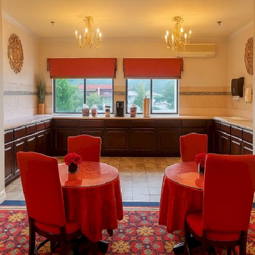 A cozy dining room with red upholstered chairs, round red-clothed tables, a patterned carpet, and a windowed counter area with warm lighting.