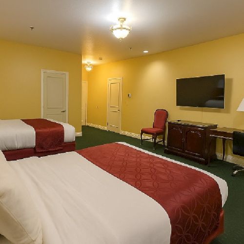A hotel room with two queen beds, yellow walls, a wall-mounted TV, a red carpet, and a desk with chairs and lamps, bright and airy.