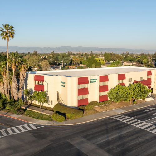 Aerial view of a small, cream-and-red building complex on a corner lot with a nearby road and blue sky.