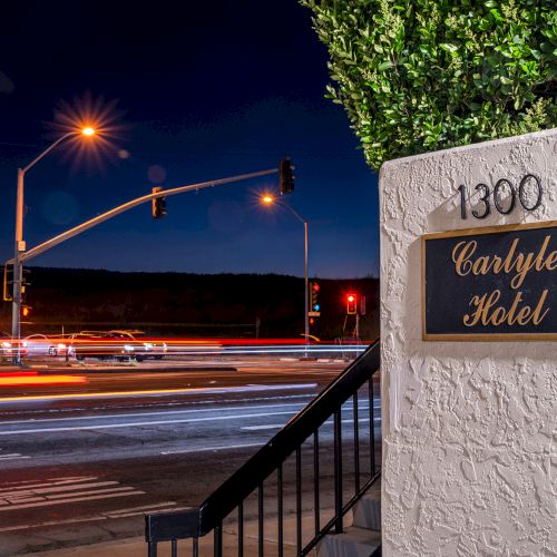 Night street scene outside a hotel entrance, light trails on the road, a white stucco wall with a sign, calm evening vibes.