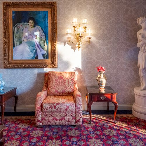 A vintage living room with ornate furniture, a gilded mirror, portraits, floral wallpaper, and a small child playing on a pink rug, end.