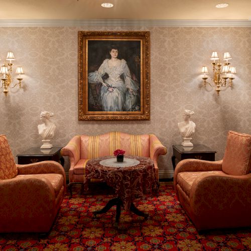 A classic, elegant living room with ornate walls, a red velvet sofa set, a framed portrait, and warm chandelier lighting.