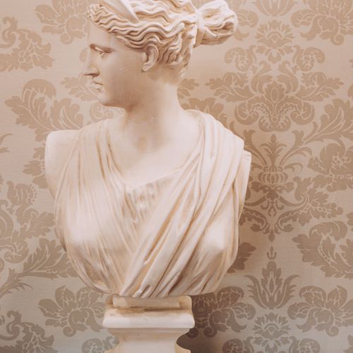 A marble bust of a classical woman with an ornate hairstyle, set on a striped pedestal against a patterned beige wall.