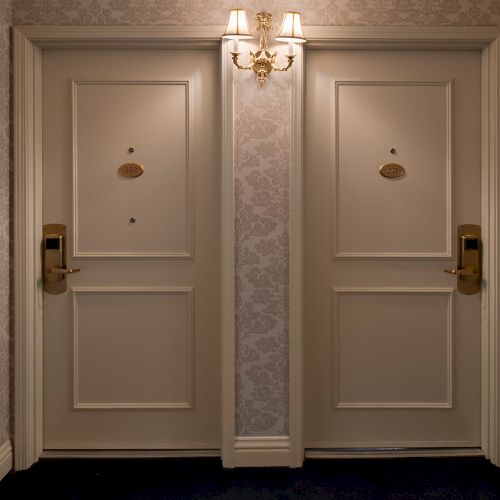 Two closed hotel doors side by side in a dim hallway, with ornate wallpaper, brass door hardware, and a small chandelier overhead.