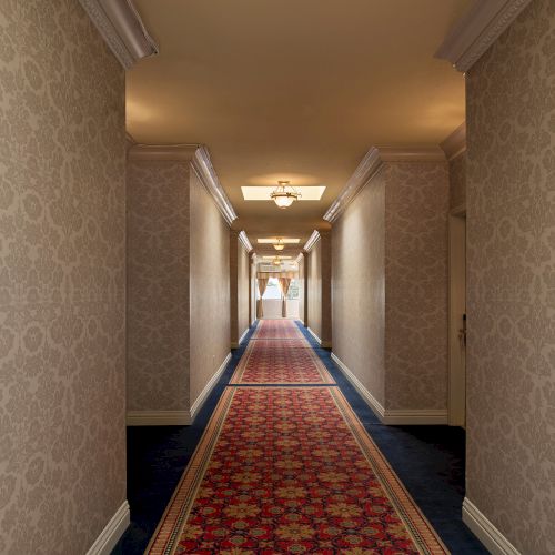 A long hotel hallway with patterned wallpaper, a red carpet runner, walls lined with doors, and soft ceiling lights fading into the distance.
