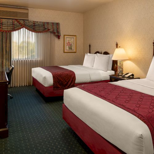 Twin beds in a hotel room with a bedside table, chair, desk, and warm lighting, topped with red bed runners.