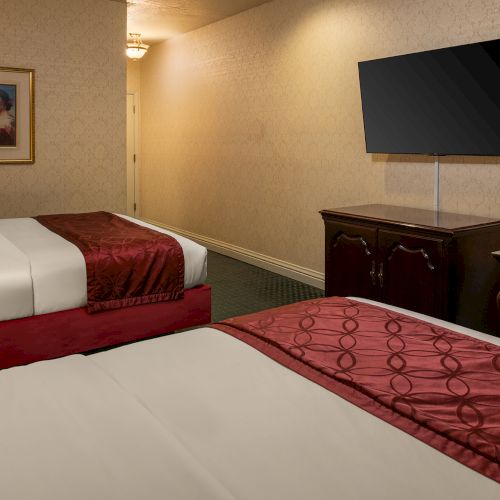 A cozy hotel room with two double beds, a desk, flat-screen TV, warm lighting, and a small dresser.