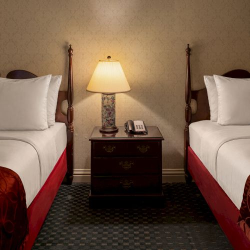 Two bedside lamps between two single beds in a dim hotel room, with a small table and two chairs, warm lighting.