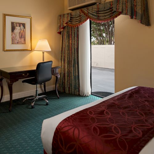 A small hotel bedroom with two single beds, a nightstand, desk, chair, and a window with curtains; vintage decor and warm lighting.