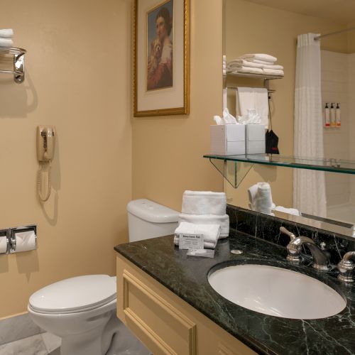 A small bathroom with beige tiled walls, a pedestal sink, and a mirror cabinet above. A towel rack, trash can, and a few toiletries on shelves.