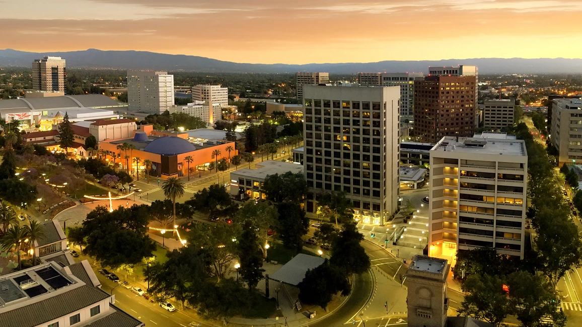 A cityscape at dusk with tall office buildings, streets, trees, and a circular park area, all illuminated by streetlights and warm sunset glow.
