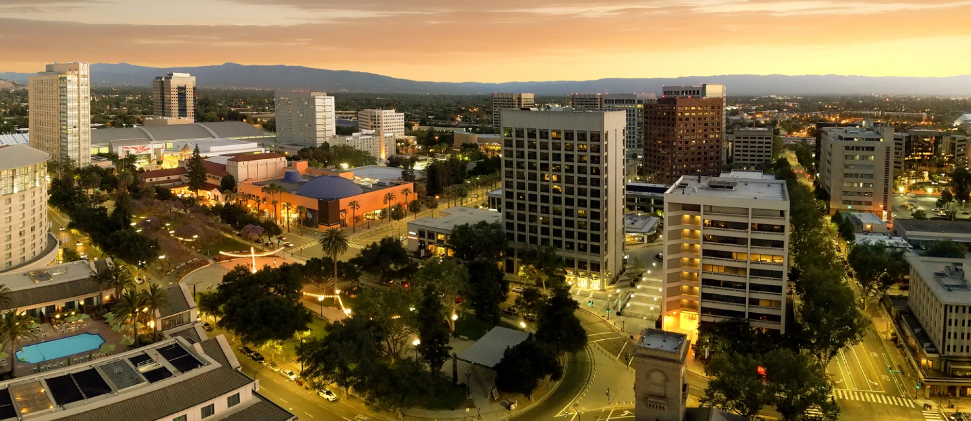 A cityscape at dusk with tall office buildings, streets, trees, and a circular park area, all illuminated by streetlights and warm sunset glow.