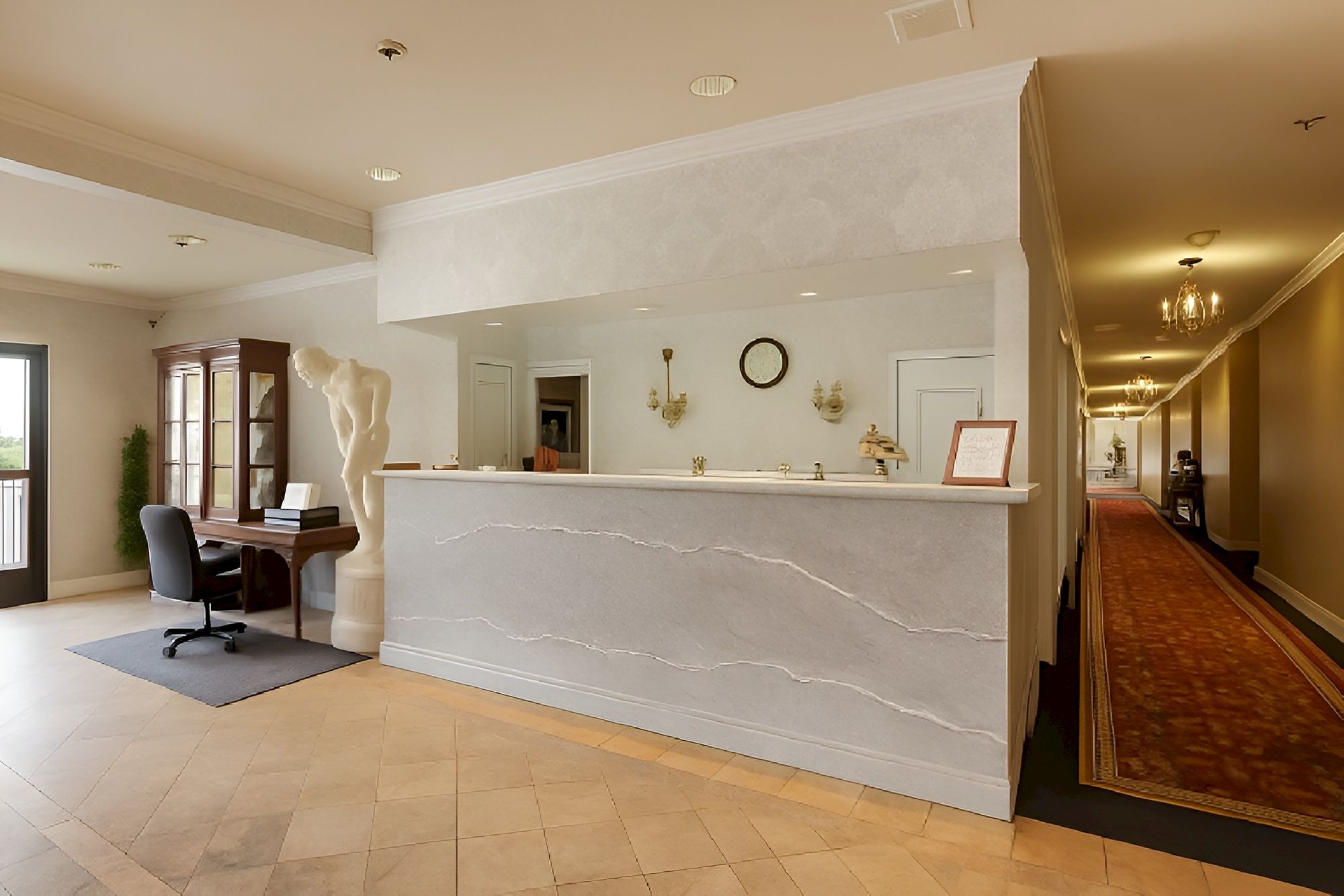 A hotel reception area with a light marble counter, a small office chair, a piano in the corner, and a hallway lined with a red carpet and wall lights.