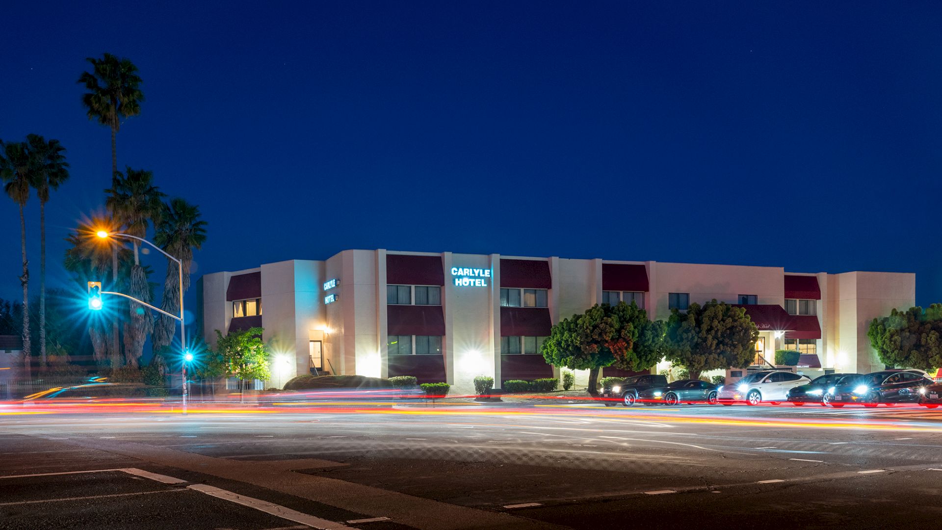 A low-rise hotel at night with lit windows, palm trees, and a street intersection showing long light trails from passing cars.