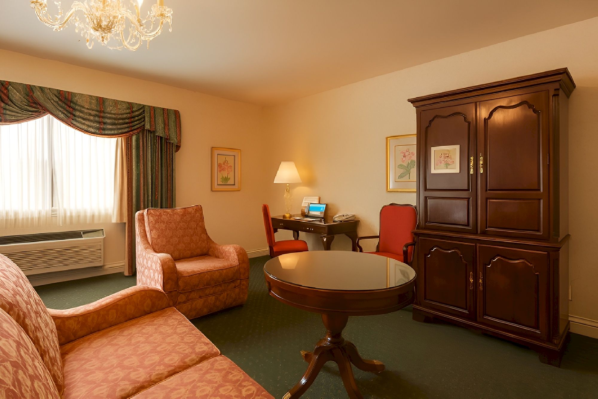 A cozy hotel lobby or parlor with pink patterned sofas, a round coffee table, a dark wooden armoire, a desk with a chair, and warm lighting.