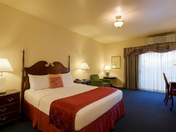 A hotel room with a large bed, two bedside lamps, a seating area, and a desk near a window with heavy drapes.