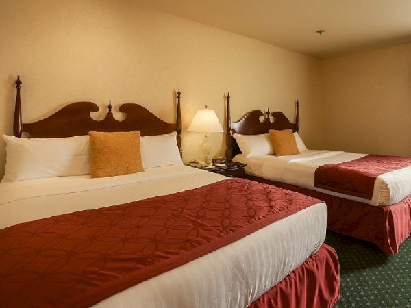 Two double beds with white linens, red bed runners, and brown headboards in a beige hotel room; a nightstand lamp sits between them, green carpet.