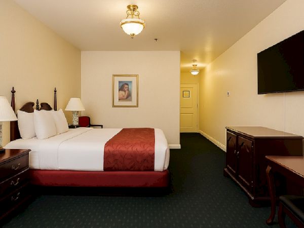 A hotel room with two large beds, white linens, red runners, nightstands with lamps, a wall-mounted TV, a dresser, a desk, and a framed picture on the far wall.