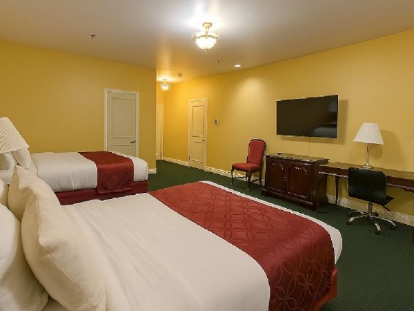 A hotel room with two double beds, yellow walls, green carpet, a desk with chair, wall-mounted TV, and lamps. The room looks tidy and bright.