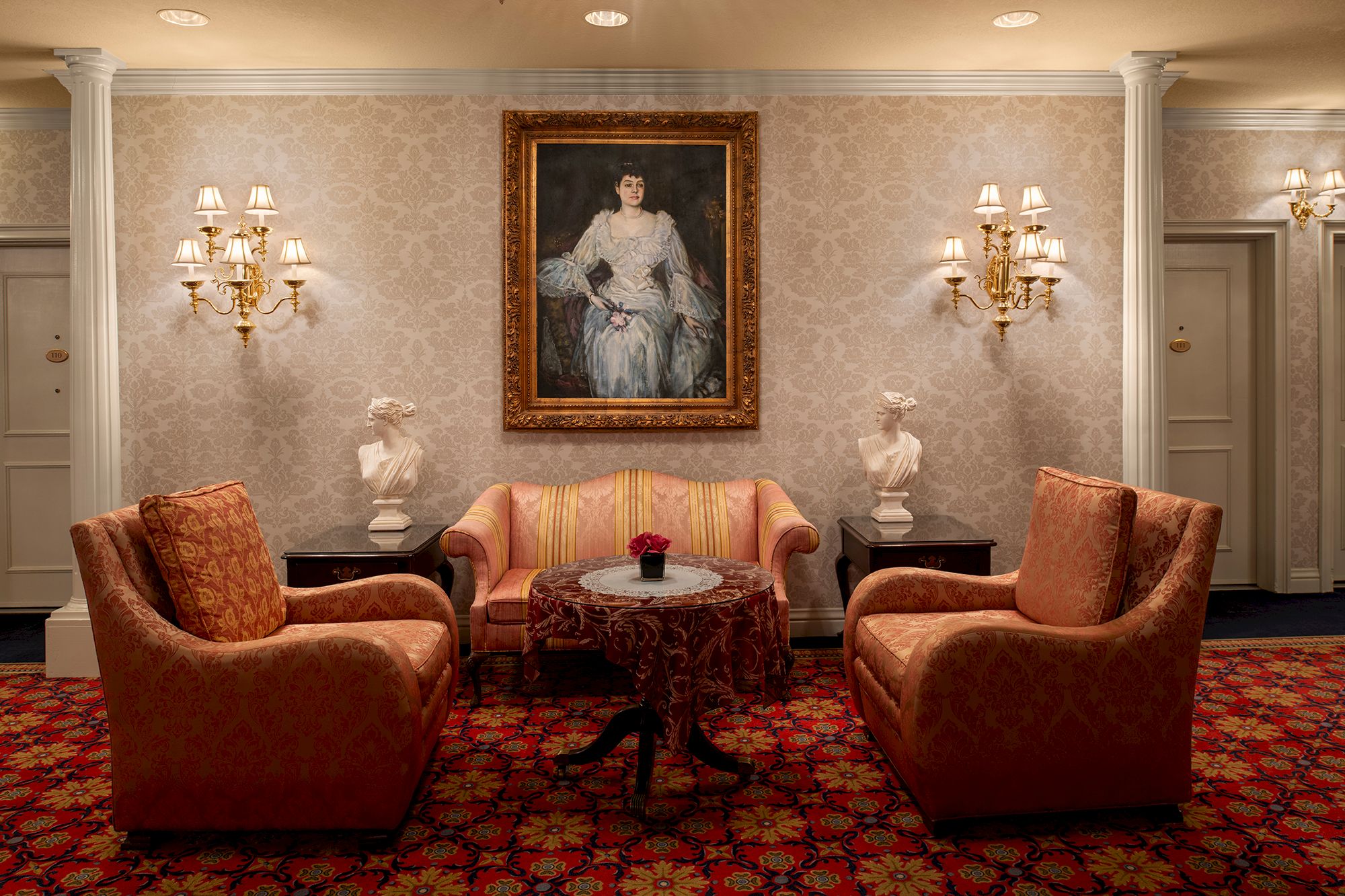 A formal hotel lobby or sitting area with four orange upholstered chairs around a round table, a portrait on the wall, and ornate lamps.