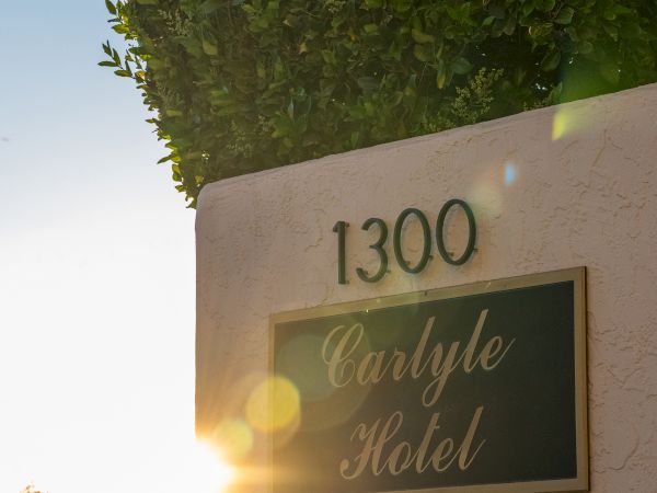 A sign shows &ldquo;1300 Carlyle Hotel&rdquo; on a sunlit wall with a hedge above, lens flare adding warm glow and a calm outdoor scene. ending sentence.