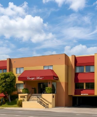 A two-story orange hotel with red awnings, manicured trees, a ramp/steps entrance, and a blue sky with fluffy clouds.