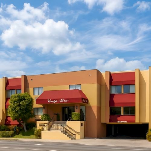 A two-story orange hotel with red awnings, manicured trees, a ramp/steps entrance, and a blue sky with fluffy clouds.