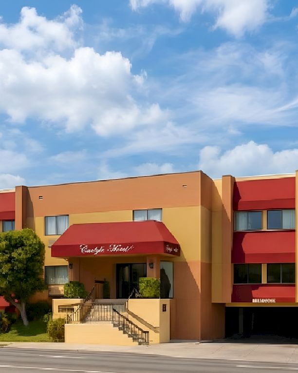 A two-story orange hotel with red awnings, manicured trees, a ramp/steps entrance, and a blue sky with fluffy clouds.