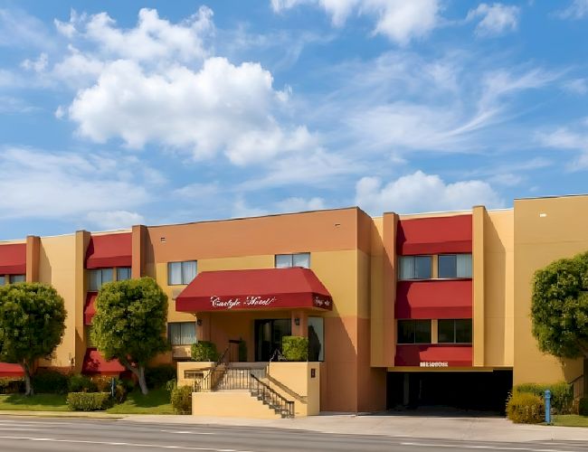 A two-story orange hotel with red awnings, manicured trees, a ramp/steps entrance, and a blue sky with fluffy clouds.