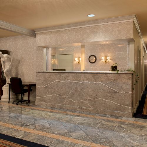 A marble hotel lobby with a front desk, a wooden cabinet, a chair, and ornate lighting, hallway to the right, polished floor, calm ambience.