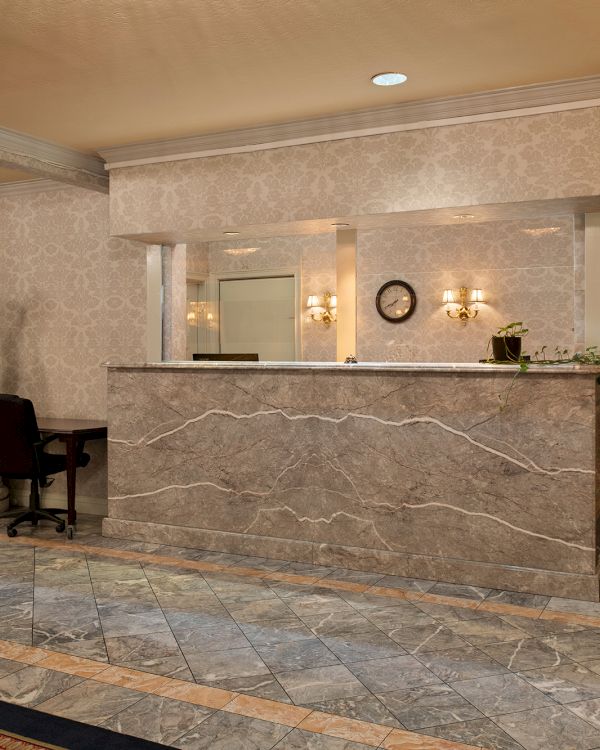 A marble hotel lobby with a front desk, a wooden cabinet, a chair, and ornate lighting, hallway to the right, polished floor, calm ambience.