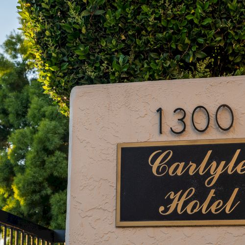 A beige stucco wall with the number 1300 above a black and gold plaque that reads &ldquo;Carlyle Hotel,&rdquo; with greenery and a fence nearby.