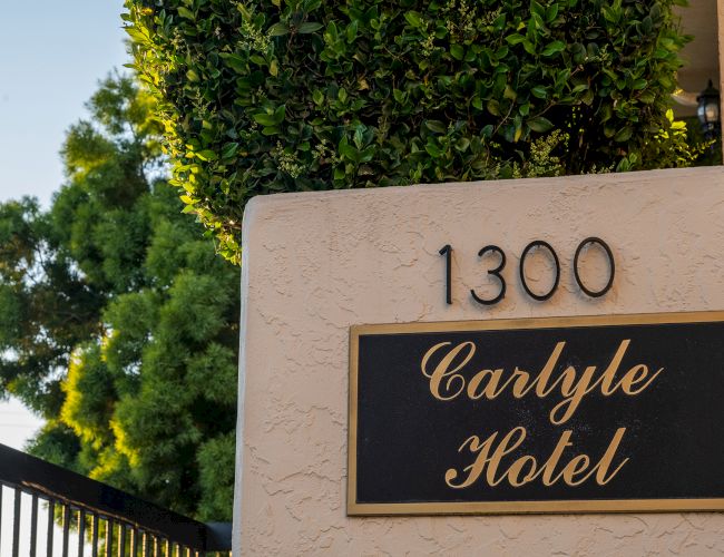 A beige stucco wall with the number 1300 above a black and gold plaque that reads &ldquo;Carlyle Hotel,&rdquo; with greenery and a fence nearby.