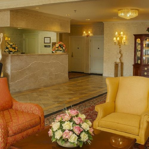 A hotel lobby scene with a marble reception desk, chic chandeliers, apricot and yellow armchairs, a wooden coffee table, and a floral centerpiece.