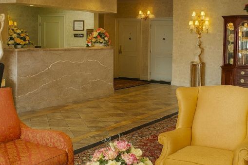 A hotel lobby scene with a marble reception desk, chic chandeliers, apricot and yellow armchairs, a wooden coffee table, and a floral centerpiece.