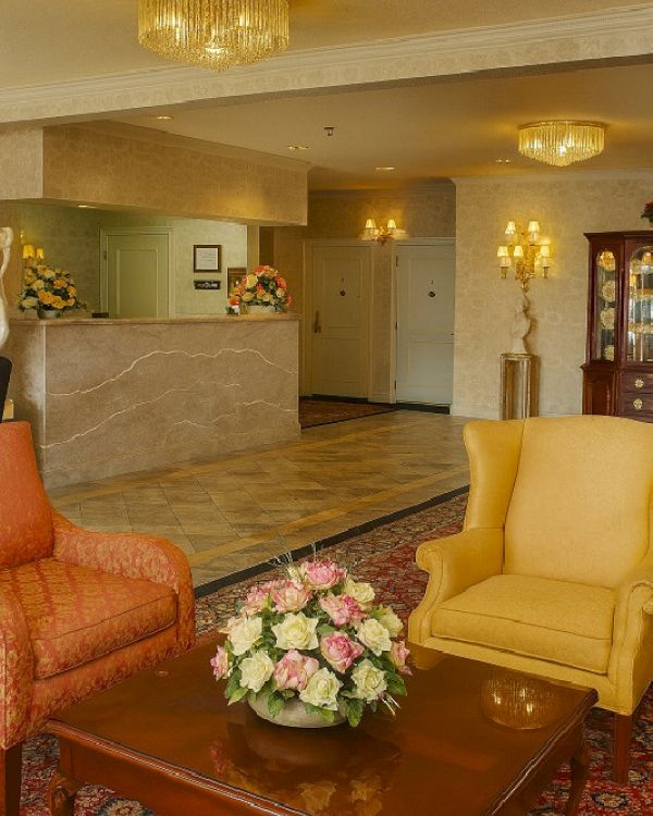 A hotel lobby scene with a marble reception desk, chic chandeliers, apricot and yellow armchairs, a wooden coffee table, and a floral centerpiece.