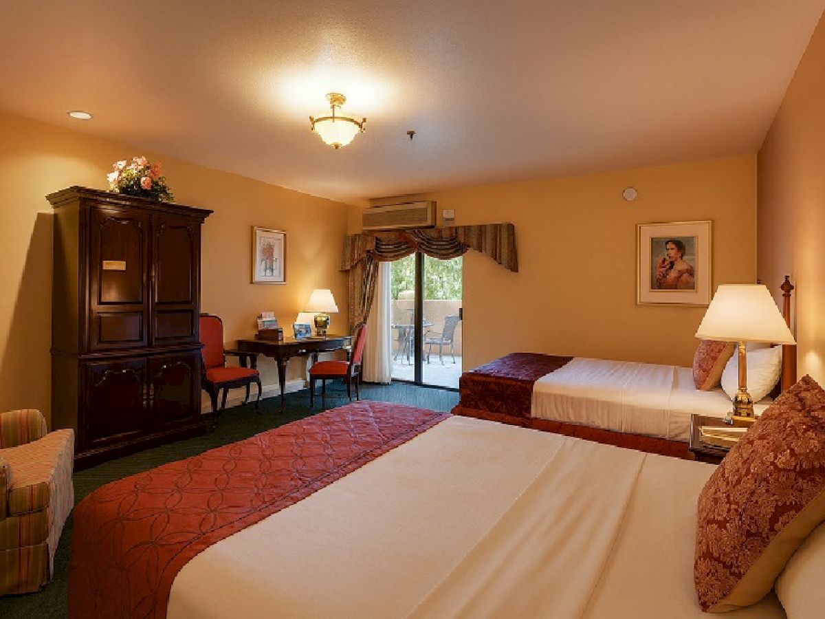 A hotel room with two queen beds, warm beige walls, a sofa, desk, armoire, and a sliding door to a balcony, soft lamps, and a red runner on each bed.