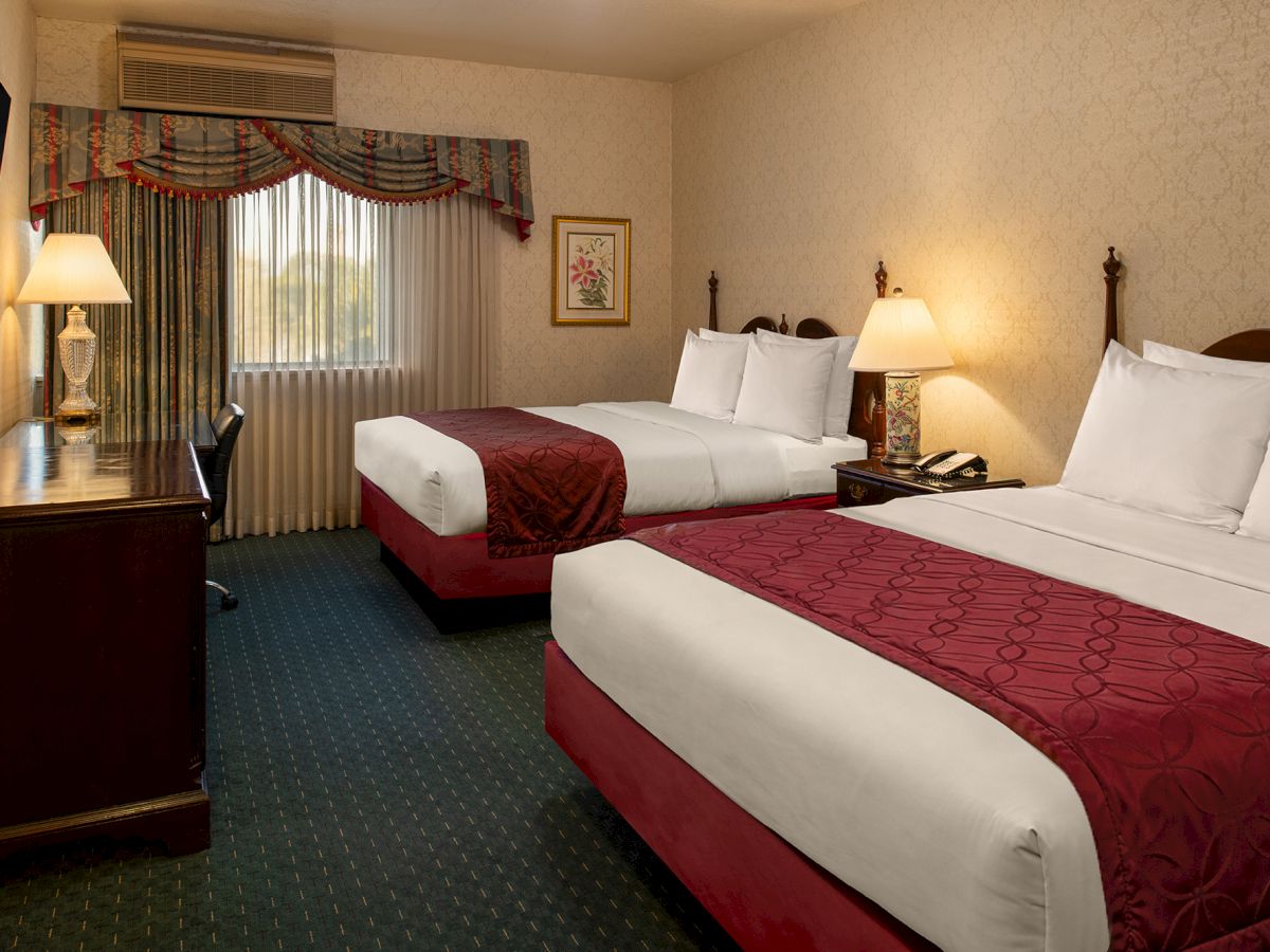 Two double beds with white linens and burgundy runners, a desk with a lamp, and a window with draped curtains in a hotel room.